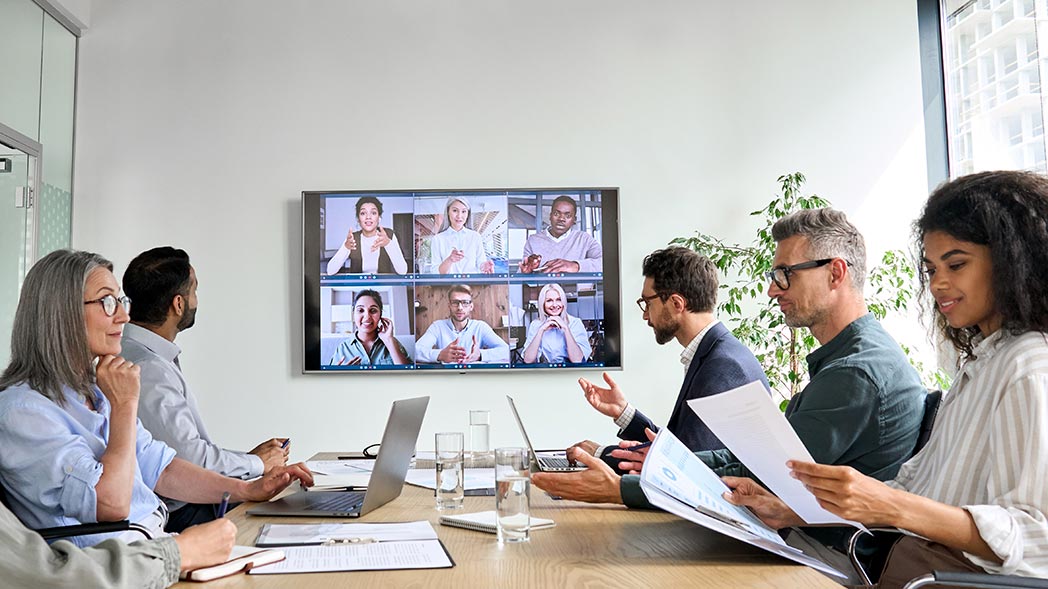 A group of people are holding a conference. Others are connected via video
